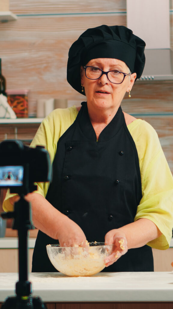 About Us 2 baker wearing bonete recording podcast traditional food recipe using flour cracked eggs influencer chef using internet technology communicating shooting social media with digital equipment About Us